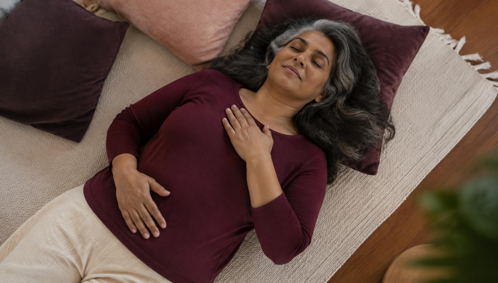 A woman of color in her mid fifties with silver-streaked hair lies on a soft rug, one hand on her heart and one on her belly.